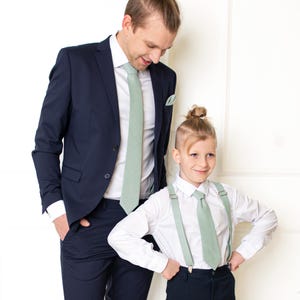 Può includere: Un uomo e un bambino che indossano cravatte verdi salvia abbinate. L'uomo indossa un abito blu scuro e il bambino indossa una camicia bianca e pantaloni neri con bretelle.