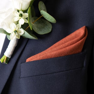 May include: A dark blue suit jacket with a white and green flower boutonniere and a rust-colored pocket square.
