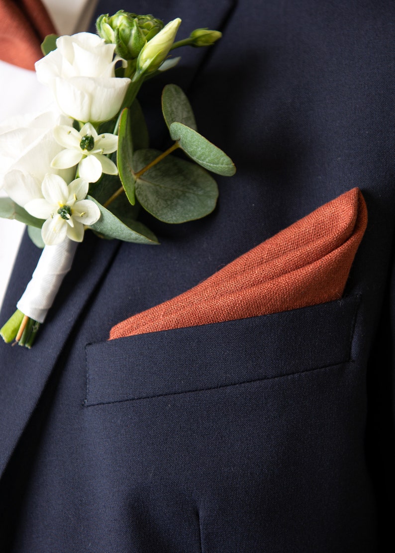 May include: A dark blue suit jacket with a white and green flower boutonniere and a rust-colored pocket square.