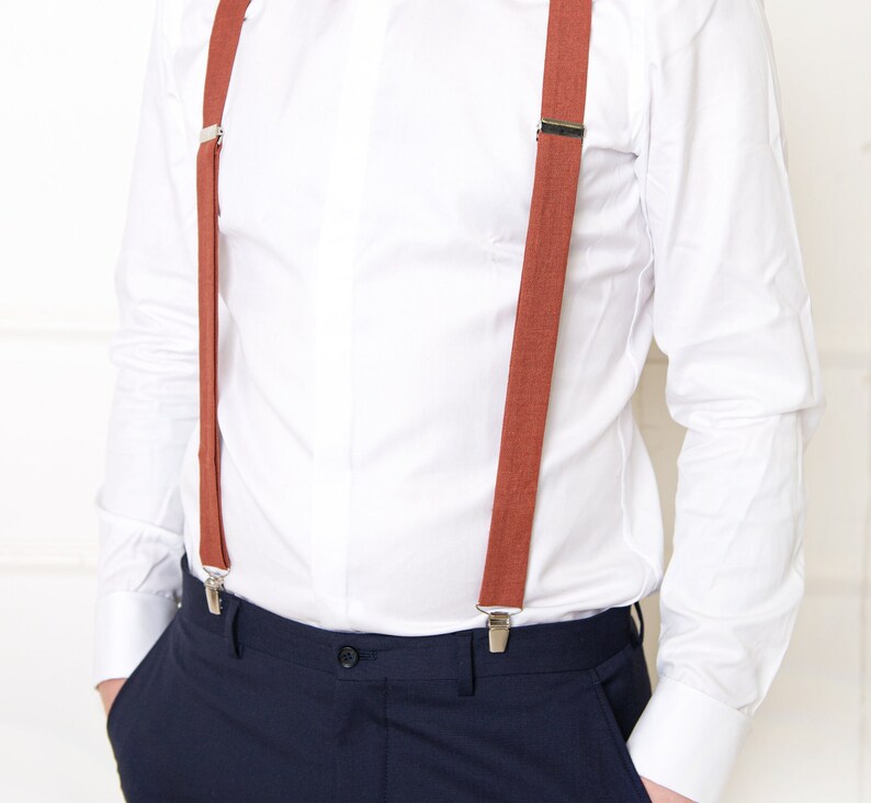 May include: A white dress shirt with a pair of brown suspenders. The suspenders have silver clips and are attached to the shirt and pants.