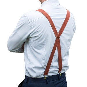 May include: A white button-down shirt with a pair of brown suspenders. The suspenders are worn over the shirt and are fastened to the pants.