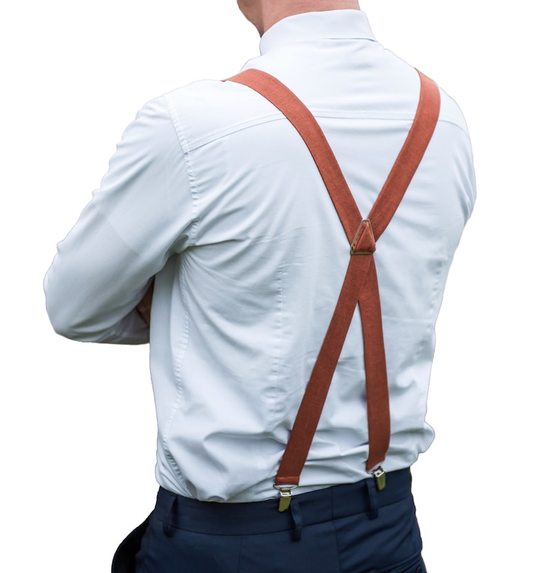 May include: A white button-down shirt with a pair of brown suspenders. The suspenders are worn over the shirt and are fastened to the pants.