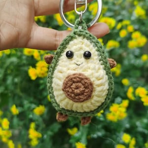Puede incluir: Un llavero de aguacate tejido a crochet con una cara sonriente, un hueso marrón y hilo verde y amarillo. El llavero está unido a un llavero plateado.