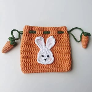 May include: Orange crocheted drawstring bag with a white bunny face and green carrot accents.