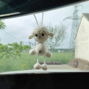 Könnte beinhalten: Weißes gehäkeltes Lamm, das an einem Rückspiegel in einem Auto hängt. Das Lamm hat schwarze Augen und einen schwarzen gestickten Mund.