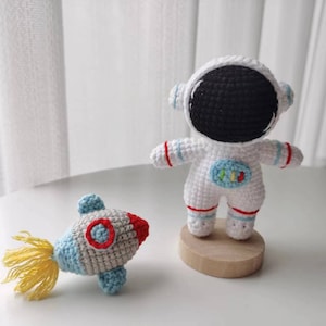 May include: A white crocheted astronaut toy with a black face and red and blue accents stands on a wooden base. A small crocheted rocket with a yellow tail and red, blue, and gray accents is in the foreground.