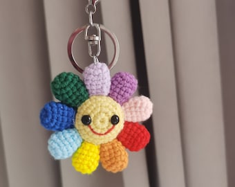 May include: A rainbow crocheted flower keychain with a smiling face. The keychain is attached to a silver key ring.