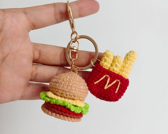 Op de afbeelding: Een gehaakte hamburger- en frietjes sleutelhanger. De hamburger is bruin met groene sla, rode tomaat en een gele kaasschijf. De frietjes zijn geel en zitten in een rode container met een witte "M" aan de voorkant.