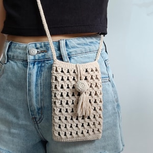 Peut inclure: Un sac de téléphone en crochet beige avec une longue bandoulière. Le sac présente un motif ajouré géométrique et une décoration en forme de pompon et de fleur. Le sac est porté à l'épaule.