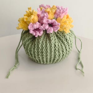 May include: A crocheted drawstring bag in light green, adorned with yellow and pink crocheted flowers. The bag has a rounded shape and is closed with a matching green drawstring. The flowers are clustered on top, creating a decorative floral design.