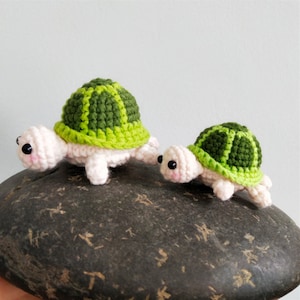 May include: Two crocheted turtle toys, one larger than the other, with green and white shells and pink bodies. The turtles are sitting on a dark grey textured surface.