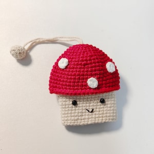 May include: A crocheted mushroom keychain with a red cap and white spots. The mushroom has a white stem with a smiling face.