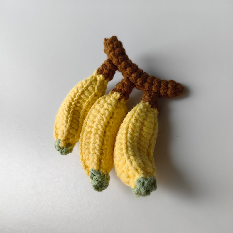 May include: A bunch of three crocheted yellow bananas with brown stems. The bananas are made with a soft yarn and have a realistic appearance.