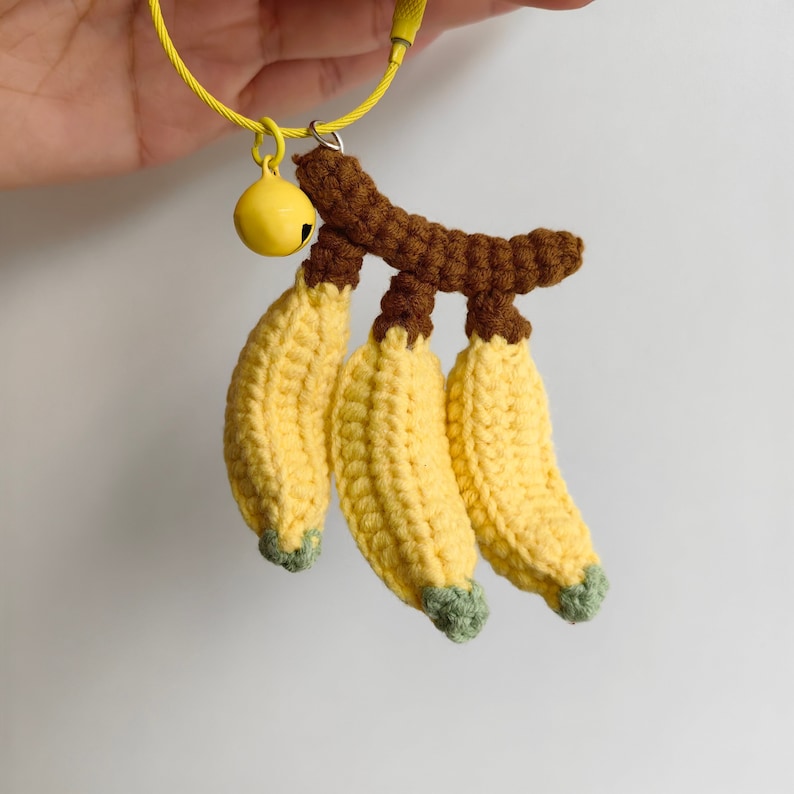 May include: A crocheted banana keychain with a brown stem and a yellow bell charm. The bananas are yellow with green tips.