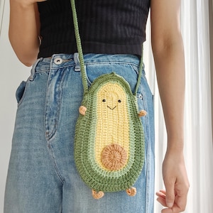 May include: A crocheted avocado-shaped crossbody bag with a smiling face and a brown seed. The bag is green and yellow and has a strap.