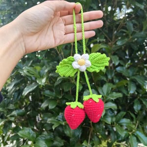 以下が含まれることがあります： 白い花と緑の葉が付いた、かぎ針編みで作ったイチゴのチャーム。チャームは緑色の紐に繋がっています。