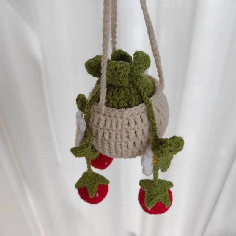 May include: A crocheted hanging planter with a green plant and three red strawberries hanging from it. The planter is beige and the strawberries are attached to green stems with white flowers.