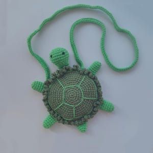 Puede incluir: Un bolso de tortuga de crochet verde con una larga correa verde. La tortuga tiene un caparazón detallado y ojos negros.