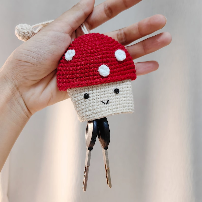 May include: A crocheted mushroom keychain with a red cap and white spots. The mushroom has a white stem with a smiling face and black eyes. The keychain is attached to a set of silver keys.