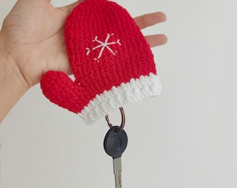 May include: Red and white crocheted mitten keychain with a snowflake design. The keychain is attached to a silver key ring with a black key.