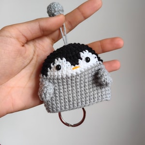Puede incluir: Un llavero de pingüino de crochet gris con ojos negros, pico naranja y vientre blanco. El pingüino está unido a un llavero plateado.