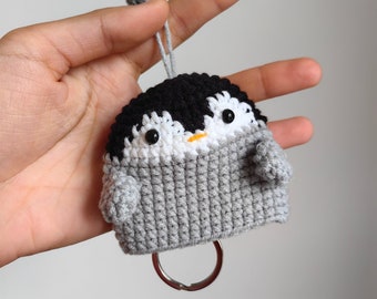 May include: A gray crocheted penguin keychain with black eyes, an orange beak, and a white belly. The penguin is attached to a silver key ring.