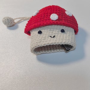 May include: A crocheted mushroom-shaped coin purse with a red top and white spots. The purse has a white crocheted body with black eyes and a smiling mouth.