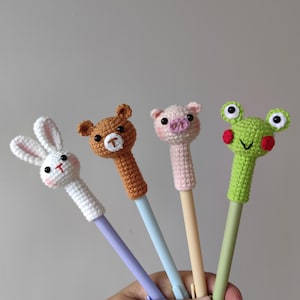 Può includere: Quattro penne all'uncinetto con decorazioni a forma di animali: un coniglio bianco, un orso marrone, un maiale rosa e una rana verde. Ogni penna ha un fusto di colore diverso. Il coniglio ha dettagli rosa, l'orso ha una T bianca, il maiale ha un muso rosa e la rana ha guance rosse.