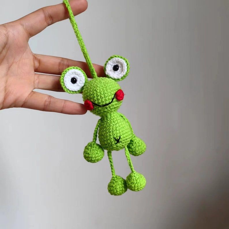 May include: A green crocheted frog toy with white eyes and red cheeks. The frog has long legs and is hanging from a string.