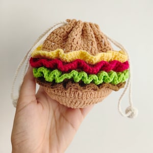 May include: A crocheted hamburger-shaped drawstring bag with a brown bun, yellow cheese, red tomato, green lettuce, and white string.