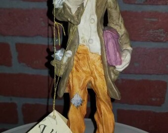 May include: A figurine of a clown holding a white dog and a purple bag. The clown is wearing a brown jacket, a white shirt, a pink bow tie, and orange pants. The figurine is on a black base with a gold label that reads "Venutti Collection".