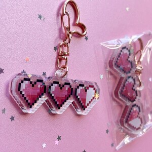 May include: A rose gold keychain featuring three connected pixel art hearts in shades of pink and white. The keychain has a heart-shaped clasp. A second identical keychain is visible inside a clear plastic bag next to the main keychain.
