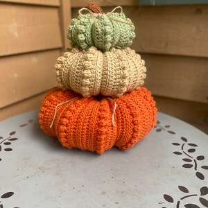 May include: A stack of three crocheted pumpkins in orange, beige, and green. The pumpkins are stacked on a white table with a decorative pattern.