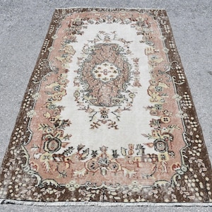 May include: Rectangular vintage rug with a central medallion design in shades of brown, beige, and cream. The rug features floral and geometric patterns, with a dark brown border. The rug is placed on a gray surface.