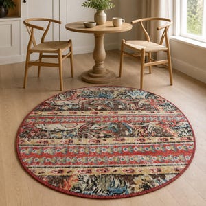 May include: A round area rug featuring a distressed, multi-coloured pattern with horizontal bands of red, beige, brown, and blue. The rug has a detailed, intricate design with a repeating motif and a solid red border edge, laid flat on a light wood floor.