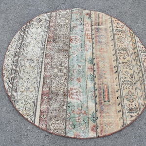 Op de afbeelding: Een rond, crèmekleurig tapijt met een veelkleurig geometrisch patroon. Het tapijt heeft een vervaagde, vintage uitstraling en is gemaakt van wol.