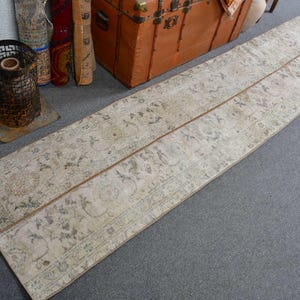 May include: A long, rectangular, vintage-style rug with a faded floral pattern in beige and muted tones. The rug is placed on a gray surface, with a brown trunk and other decorative items in the background.