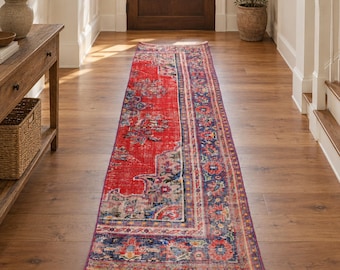 Puede incluir: Una alfombra pasillera larga y rectangular que presenta un campo central de color rojo vibrante con intrincados patrones florales y geométricos en tonos azul, crema y naranja. La alfombra tiene un borde detallado y flecos cortos.