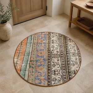 Puede incluir: Una alfombra redonda con un diseño de patchwork que presenta franjas verticales de diversos patrones. La alfombra muestra una paleta de colores naranja, azul, verde y crema, con intrincados motivos florales y geométricos en su superficie.