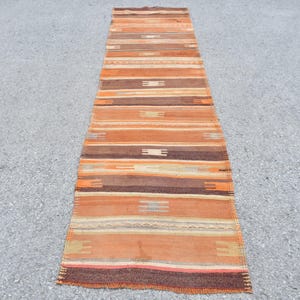 May include: A long, narrow rug with a striped pattern in shades of brown, orange, and beige. The rug has a fringe on both ends.