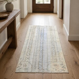 May include: A rectangular runner rug featuring a faded, distressed pattern in muted tones of beige, cream, and soft blue. The design includes intricate, worn-out geometric and floral motifs across the surface, finished with short fringe at both ends.