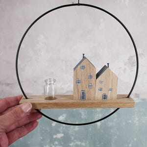 May include: A wall-hanging decoration featuring a black metal hoop framing a horizontal wooden plank. On the plank, there are two small wooden house figures with painted windows and doors, alongside a tiny, clear glass bottle vase.