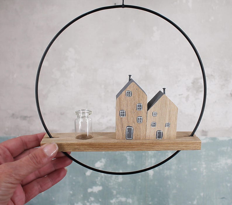 May include: A wall-hanging decoration featuring a black metal hoop framing a horizontal wooden plank. On the plank, there are two small wooden house figures with painted windows and doors, alongside a tiny, clear glass bottle vase.