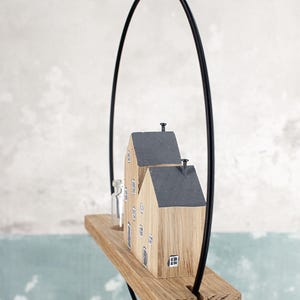 May include: A hanging wooden decoration featuring two small house-shaped blocks with dark gray roofs and painted windows, mounted on a rectangular wooden base with a clear glass bottle, all suspended within a thin black metal circular frame.