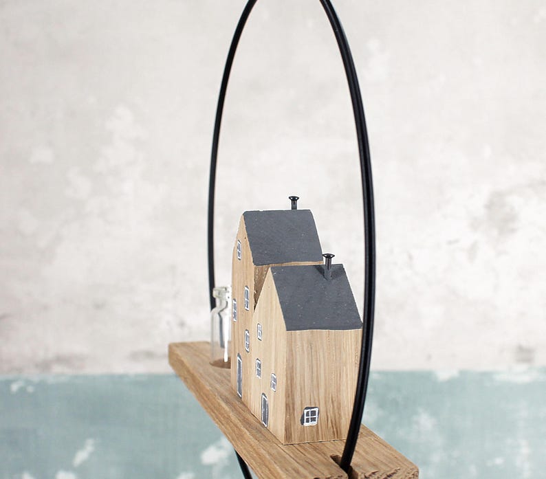 May include: A hanging wooden decoration featuring two small house-shaped blocks with dark gray roofs and painted windows, mounted on a rectangular wooden base with a clear glass bottle, all suspended within a thin black metal circular frame.