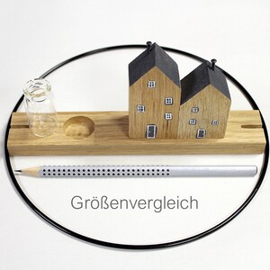 May include: A rectangular wooden base features two miniature house figures with black roofs and painted windows, a small clear glass vial, and a circular indentation. A silver pencil with a dotted pattern lies in front, with the text Gr&ouml;ssenvergleich.