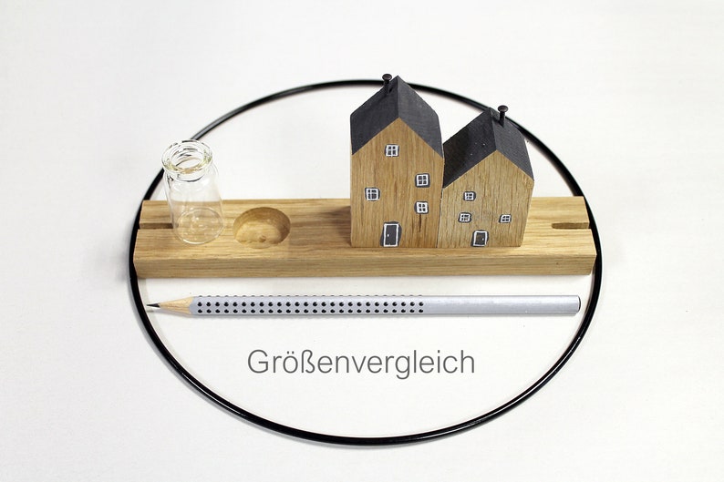 May include: A rectangular wooden base features two miniature house figures with black roofs and painted windows, a small clear glass vial, and a circular indentation. A silver pencil with a dotted pattern lies in front, with the text Gr&ouml;ssenvergleich.