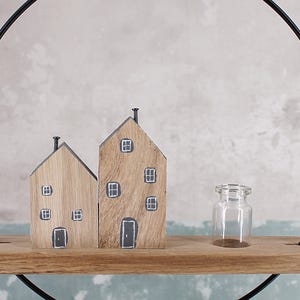 May include: A decorative display featuring two wooden house figurines with painted windows and doors, and a small clear glass bottle, all resting on a horizontal wooden shelf attached to a large black metal circular frame.