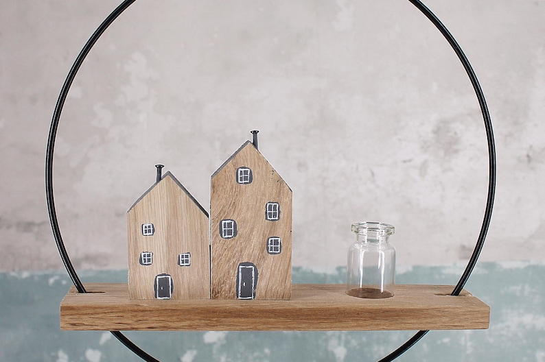 May include: A decorative display featuring two wooden house figurines with painted windows and doors, and a small clear glass bottle, all resting on a horizontal wooden shelf attached to a large black metal circular frame.