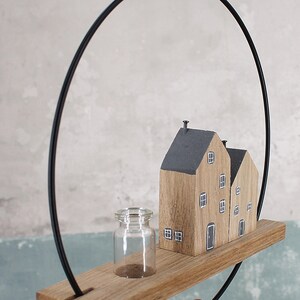 May include: A decorative wall hanging featuring a thin black metal hoop attached to a horizontal wooden plank. On the plank sits a small clear glass bottle and two miniature wooden house figures with dark gray roofs and painted white windows.